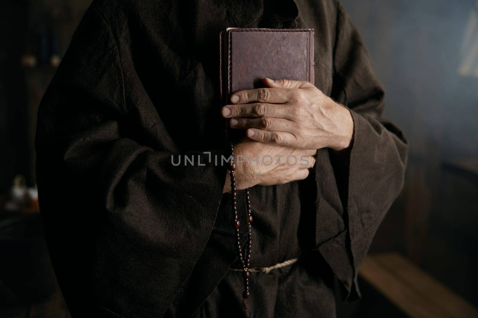 Priest wearing medieval robe clothing holding closed bible book and rosary beads in hands closeup