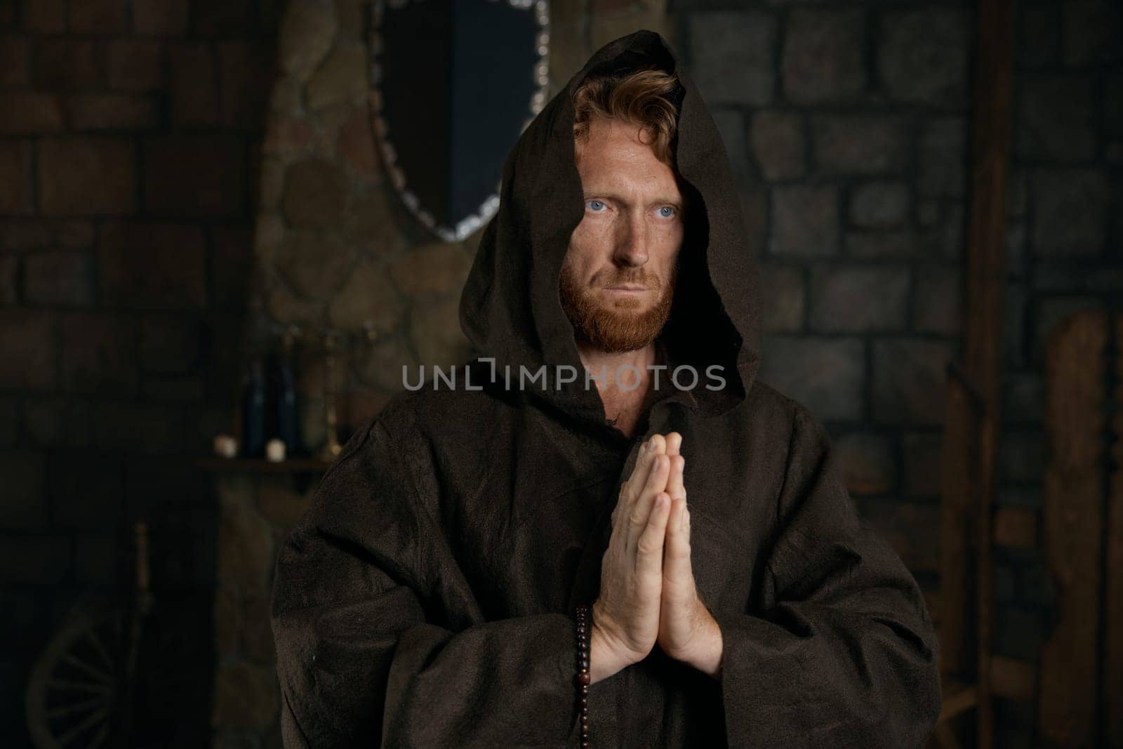 Portrait of blessed priest praying having faith believe in God. Monk wearing robe with hood holding folded hands front of chest standing over quiet dark mystical monastic background