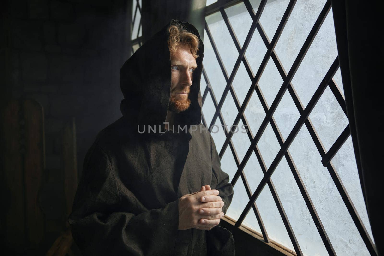 Pensive monk in medieval costume with hood looking through window of monastery. Silent prayer and appeal to God