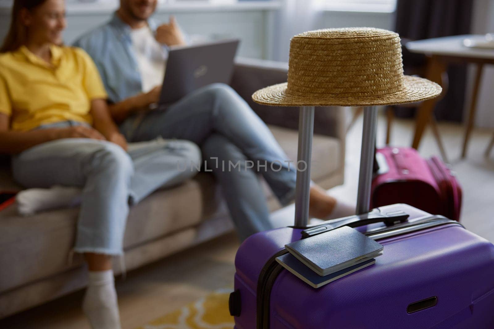 Closeup view straw hat, passport and sunglasses on travel baggage packed for vacation with happy married couple sitting and browsing internet on laptop