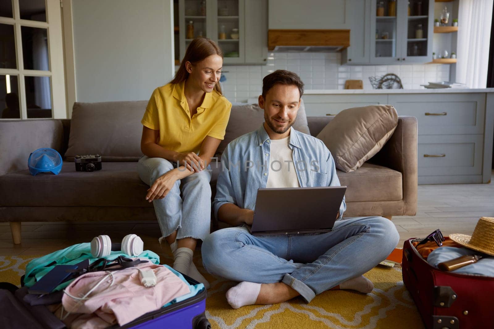Loving couple booking vacation trip choosing flight on internet via laptop while sitting in home living-room with opened travel suitcases
