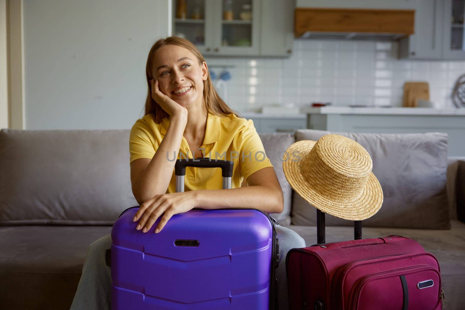 Happy woman wearing straw summer hat with travel luggage getting ready for vacation. Pretty girl tourist sitting on home sofa waiting for plane to tropical resort