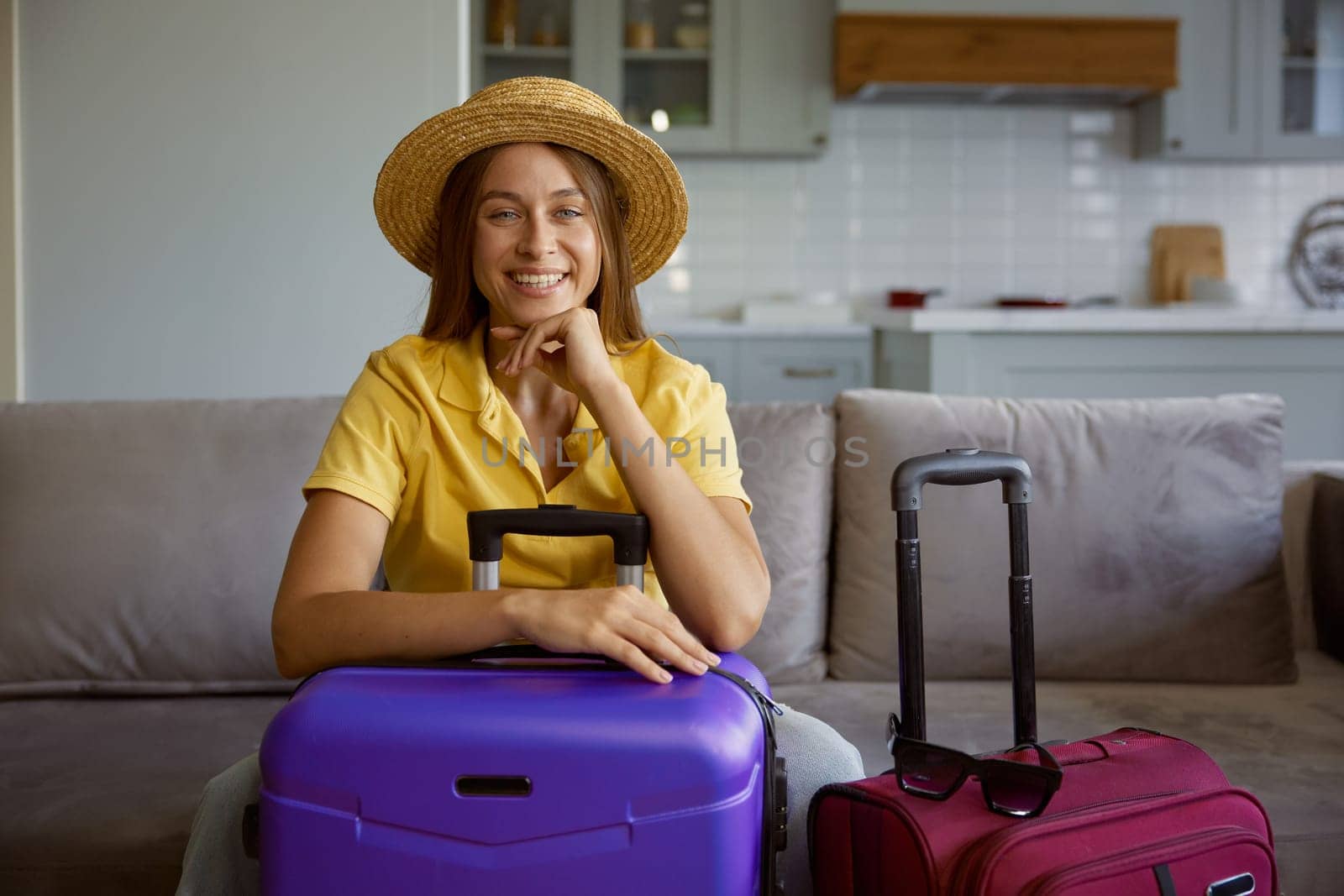 Portrait of happy woman wearing straw summer hat with travel luggage getting ready for vacation. Pretty girl tourist sitting on home sofa waiting for plane to tropical resort