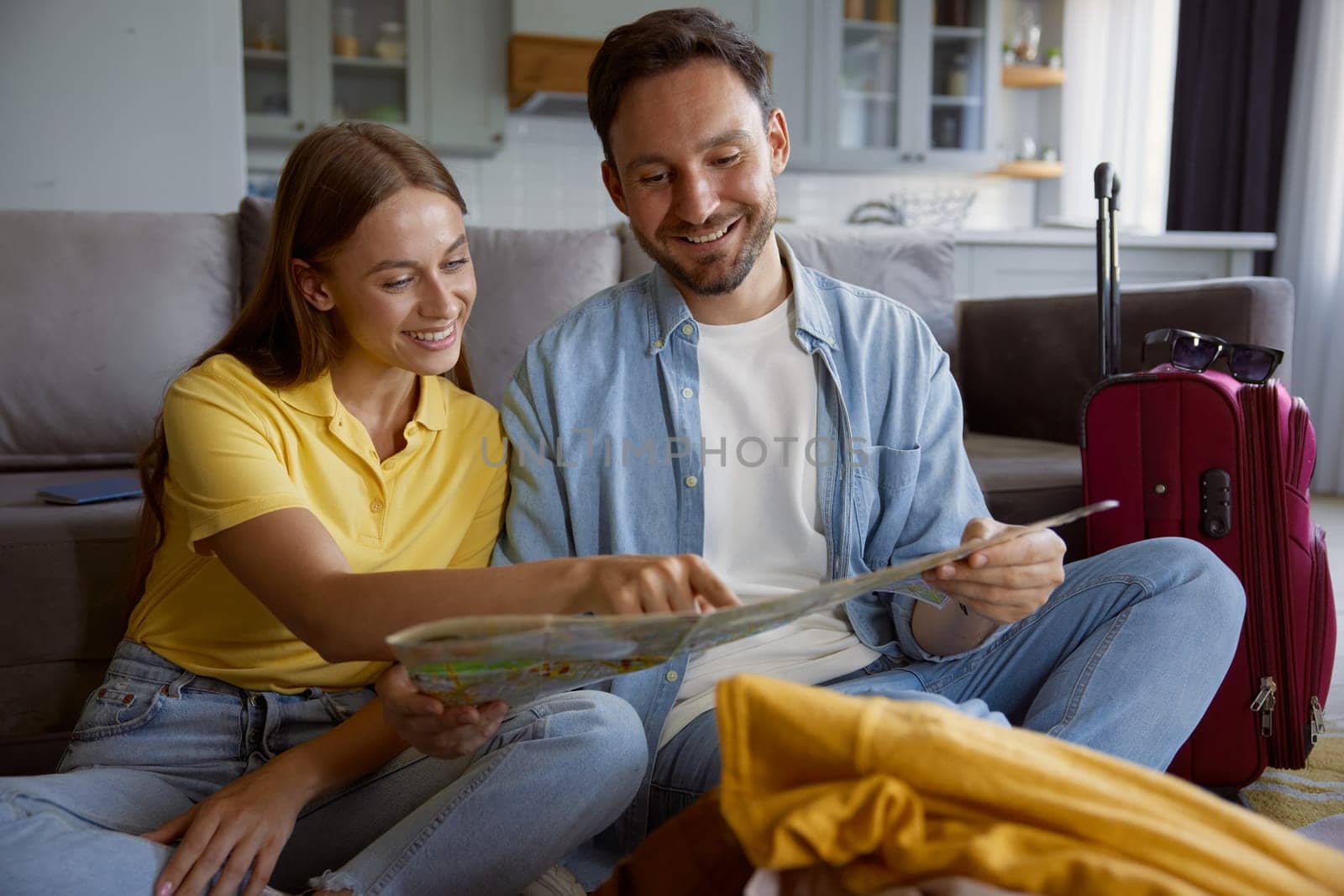Happy loving couple reading paper map planning travel vacation during summer holidays. Cheerful man and woman sitting on floor front of stack of clothing prepared for trip