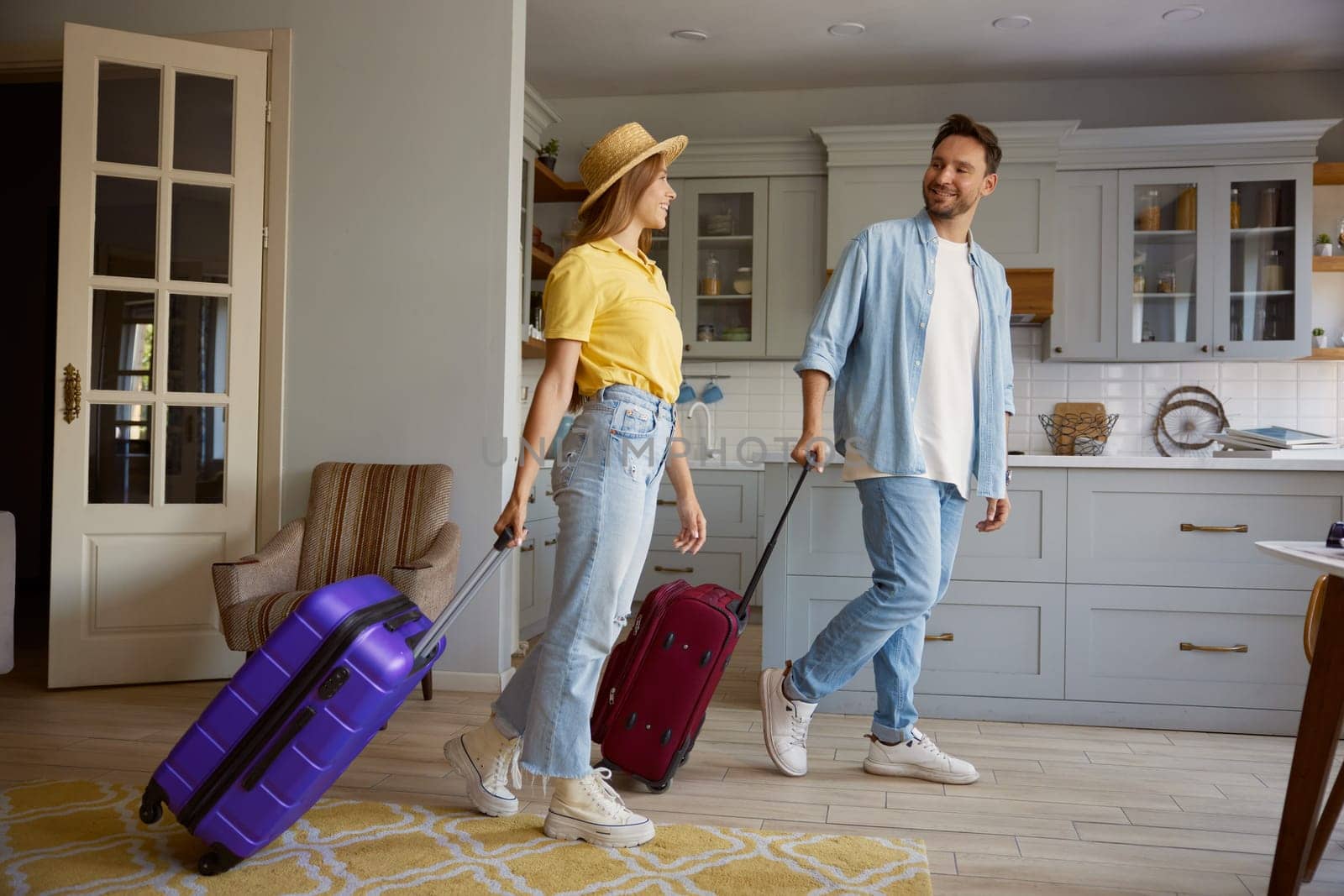 Happy smiling loving couple leaving home apartment with packed luggage ready for summer vacation trip, young man and woman feeling excitement and fun walking with suitcases