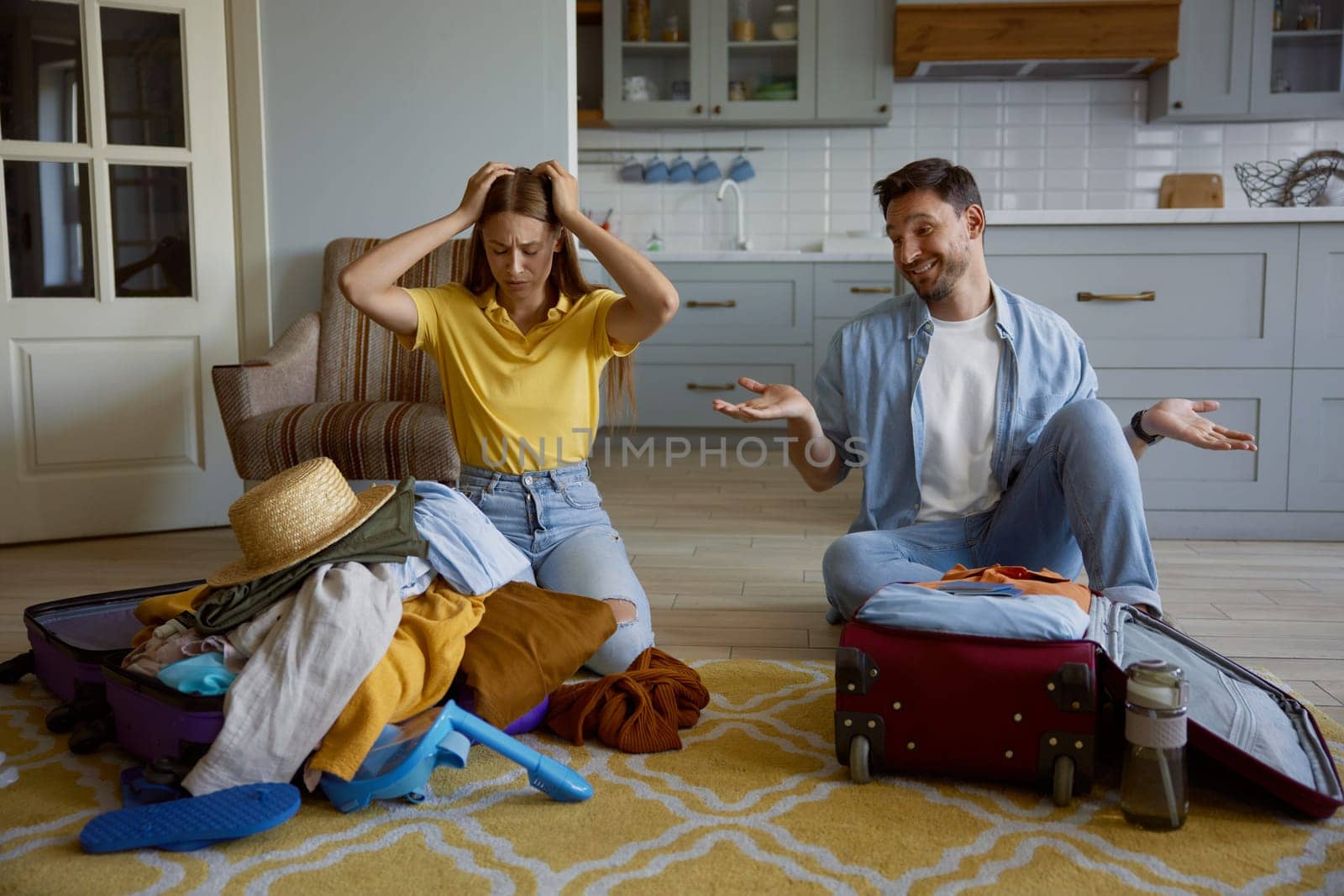 Funny married couple packing clothes for travel vacation at home. Upset woman sitting front of excess of clothes in opened suitcase and happy man folding belongings in one bag