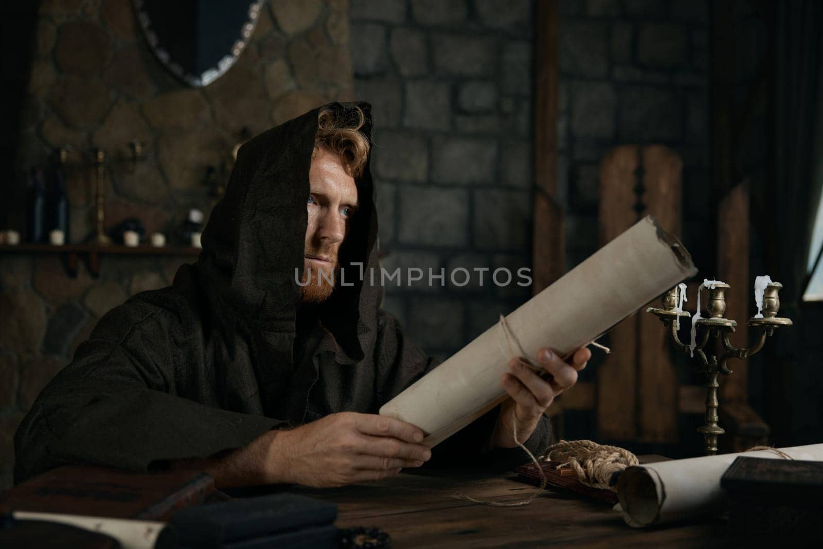 Religious man in hooded cape holding obsolete parchment scroll in hands sitting at wooden table of old mediaeval cathedral