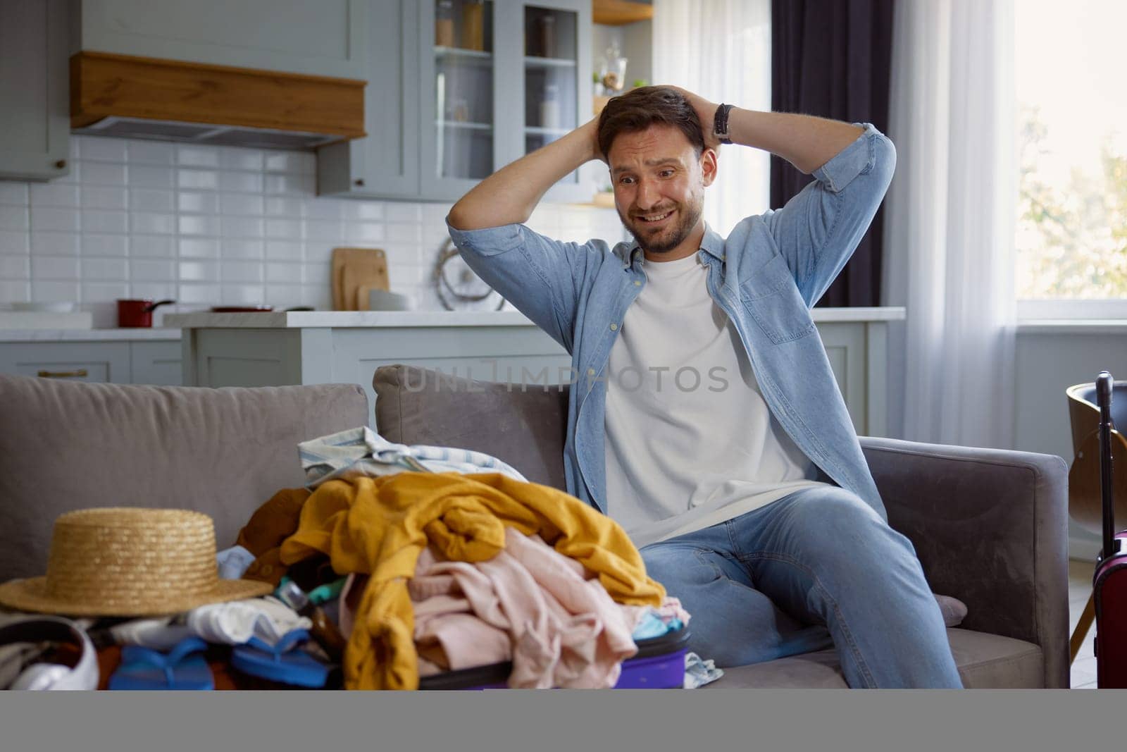 Unhappy worried man touching head with crazy emotion looking on stack of clothing in opened travel suitcase. Preparation for trip and overloaded luggage bag
