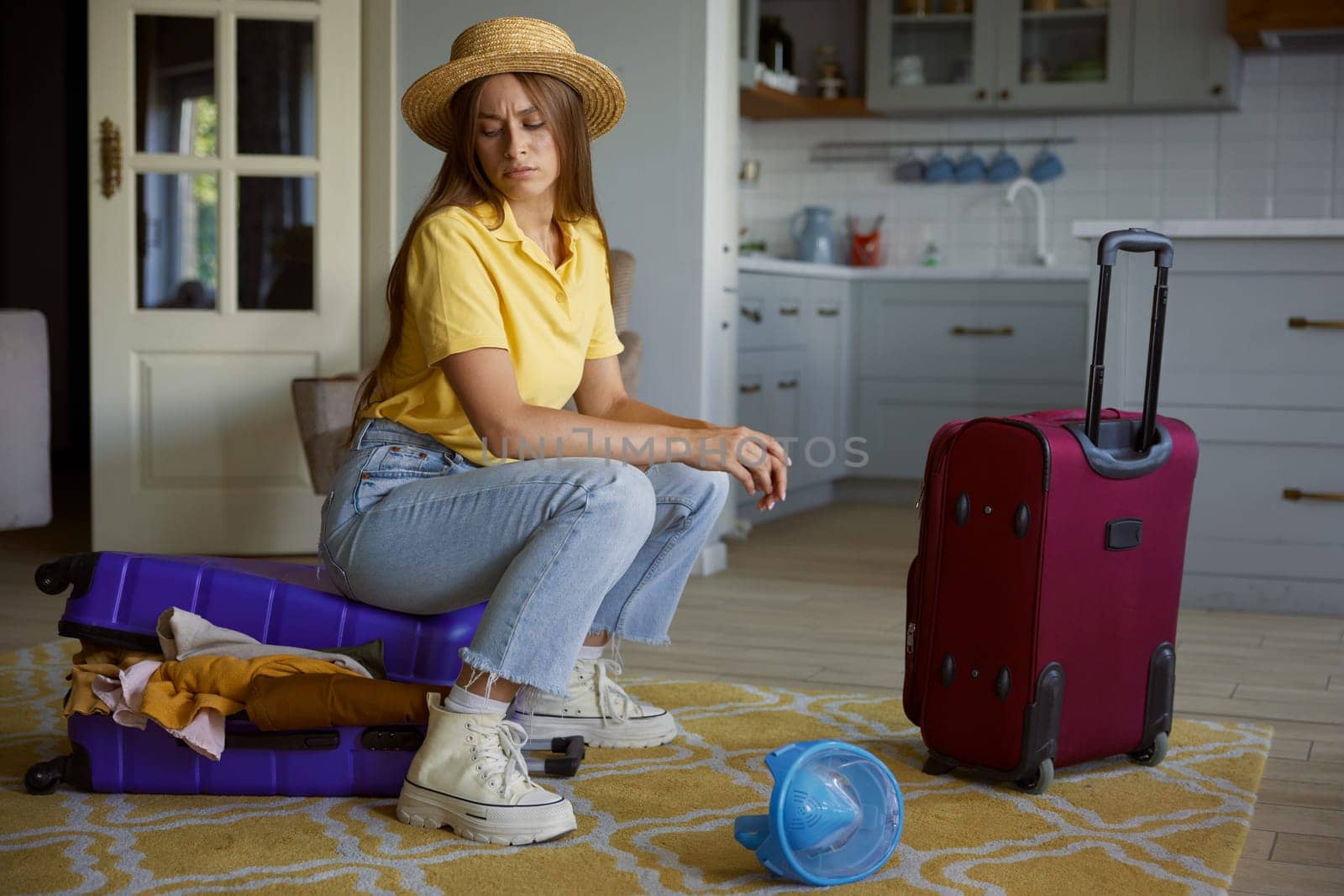 Upset woman tourist frowning face sitting on travel suitcase trying to pack lots of clothes for vacation on summer resort