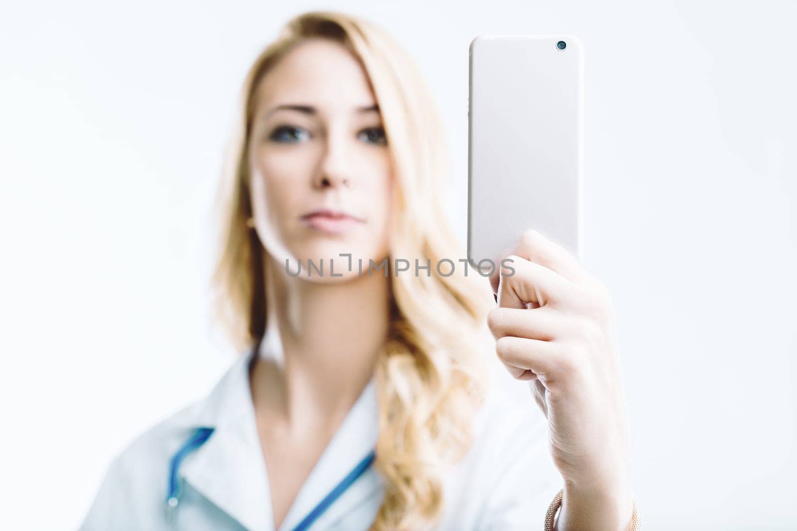 Young female doctor holding a smartphone with a blank screen, looking directly at the camera against a clean white background, exuding professionalism in her medical uniform