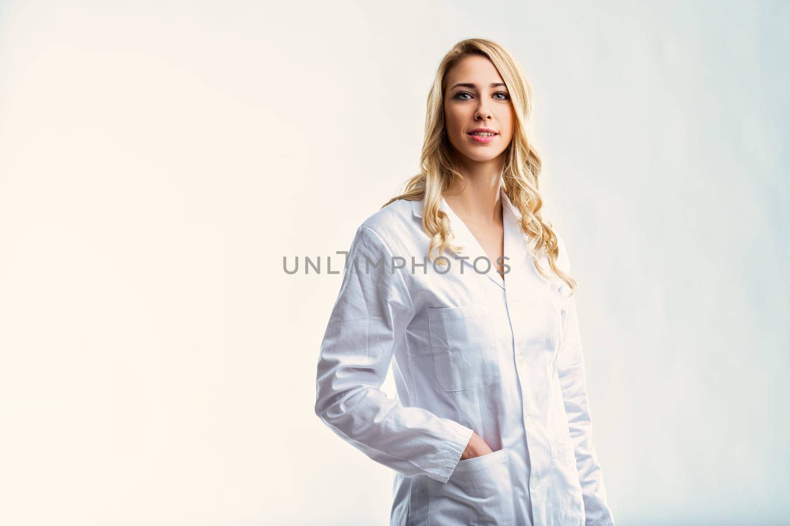 Young female doctor with long blond hair, wearing a white coat, stands confidently with her hands in her pockets against a white backdrop, exuding professionalism and expertise
