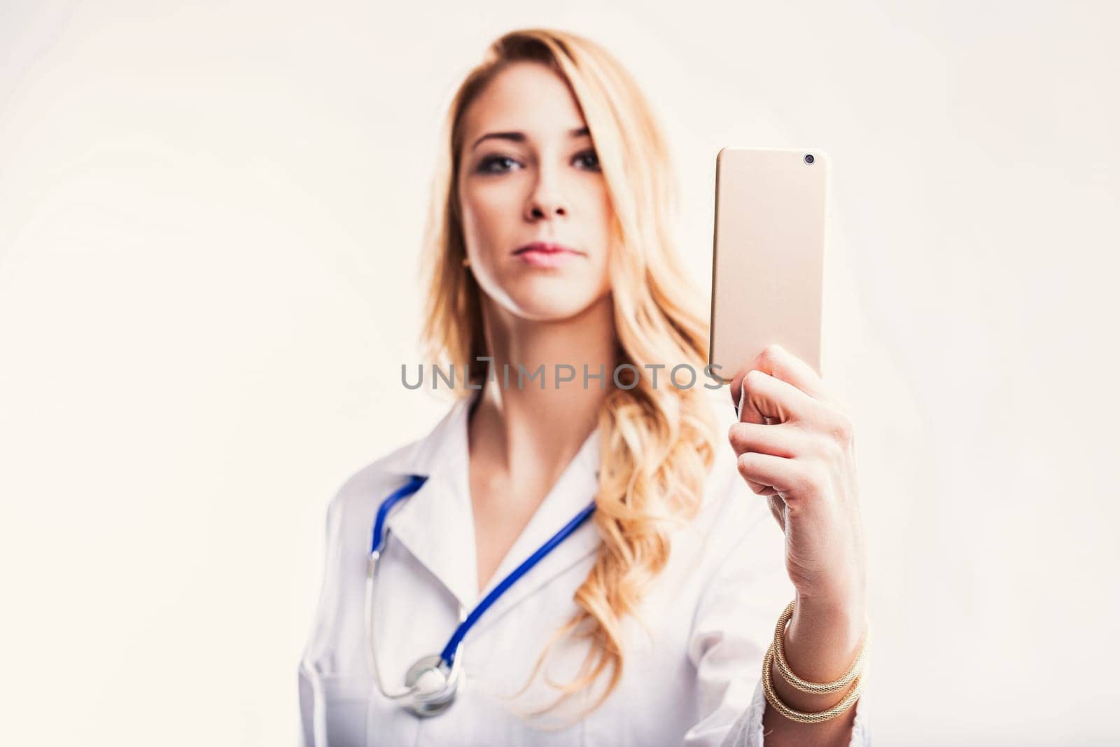 Confident young doctor wearing lab coat and stethoscope is taking a selfie with her smartphone, she has long blond hair and is looking at the camera