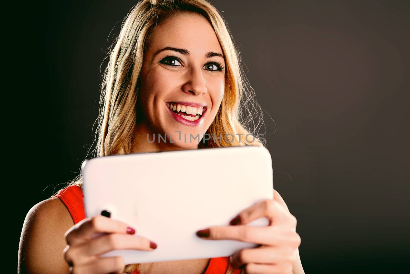 Young blonde woman with red dress holding a tablet and playing with it, smiling and looking at the camera, having fun with technology and entertainment