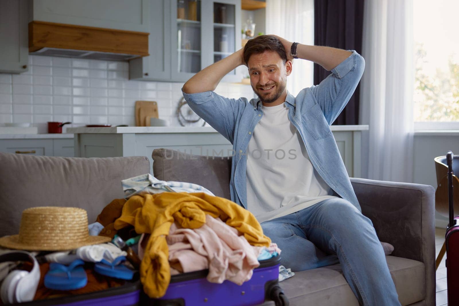 Unhappy worried man touching head with crazy emotion looking on stack of clothing in opened travel suitcase. Preparation for trip and overloaded luggage bag