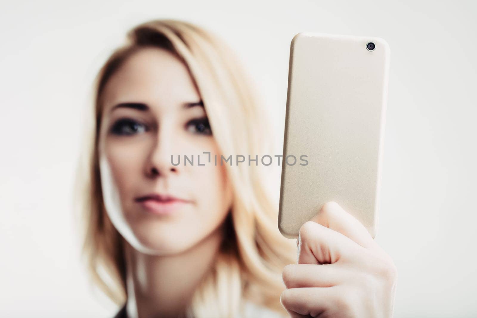 Blonde young woman is holding a smartphone in front of her face, taking a selfie, with a serious and focused expression, against a white background