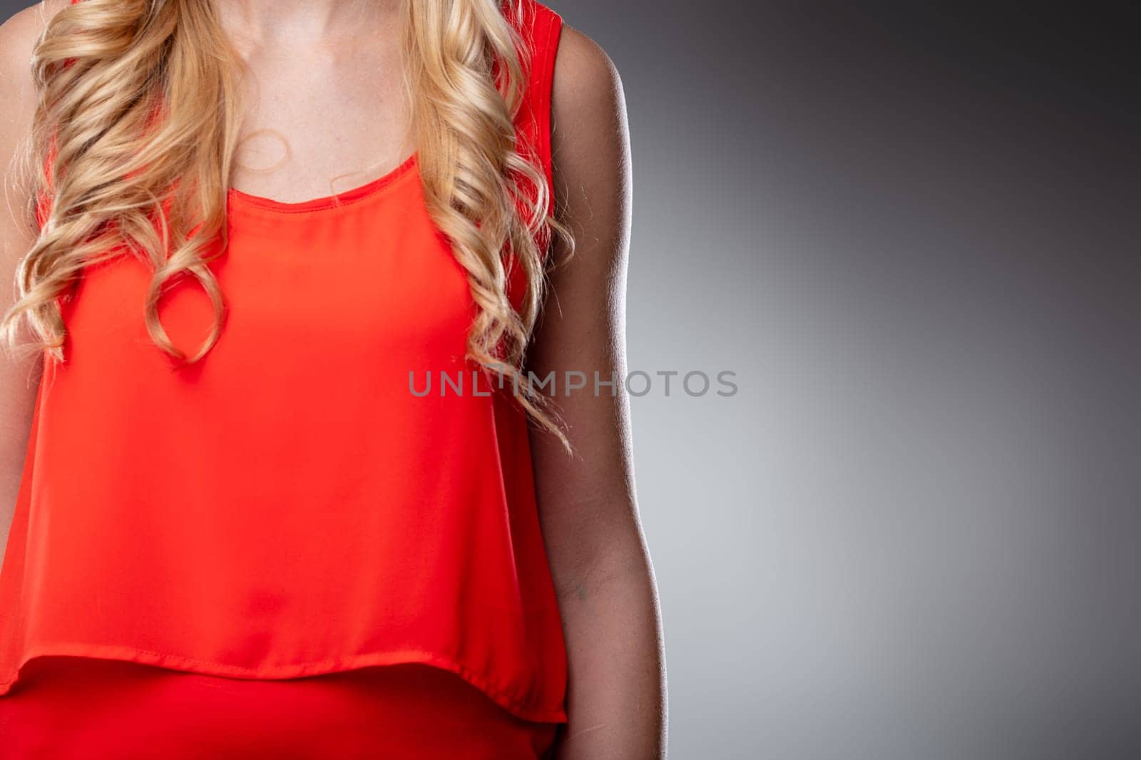 Woman in bright orange dress, dark background. We see her blonde curls, arm, but not her face. Background with ample copyspace suggests topics related to the female figure