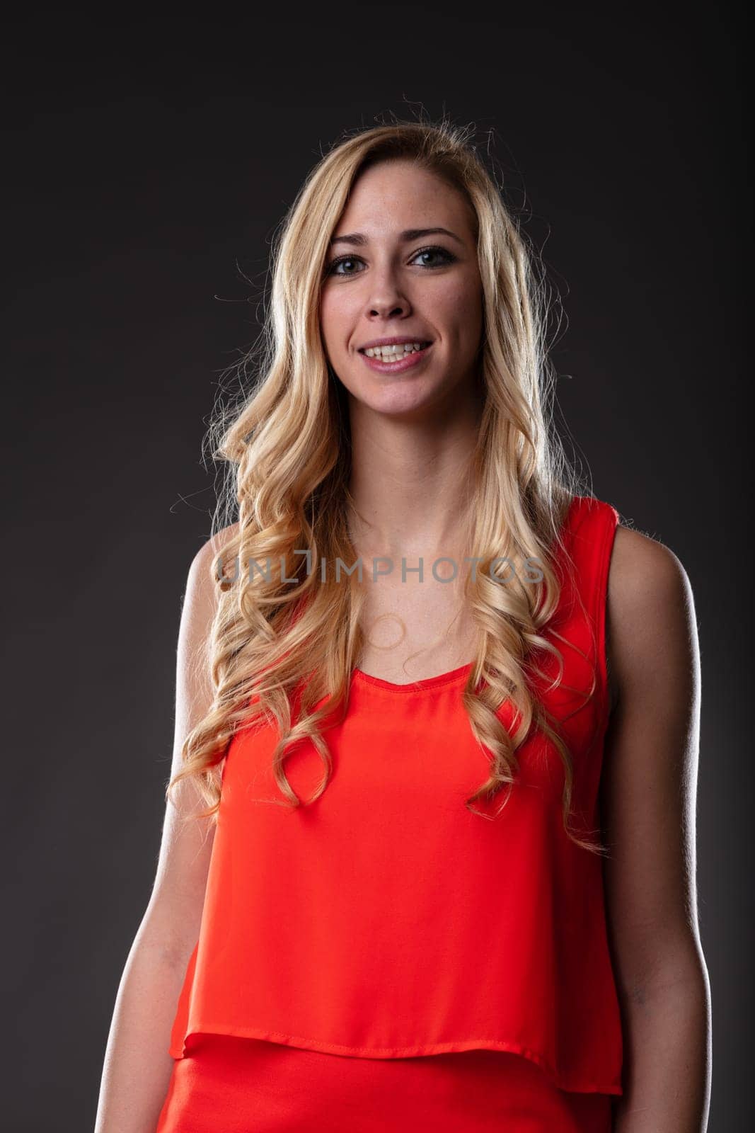 A blonde woman in an orange dress, caught in a half-length portrait. Gentle smile, relaxed arms, black backdrop, accented by light from the right