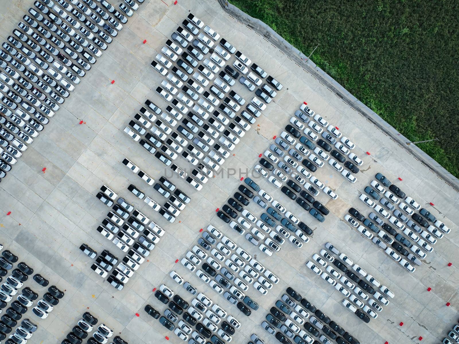 Aerial view of a factory parking lot filled with new cars adjacent to lush green grass field. Automotive industry's focus on sustainability, logistics, and zero-emission electric and hybrid vehicles.