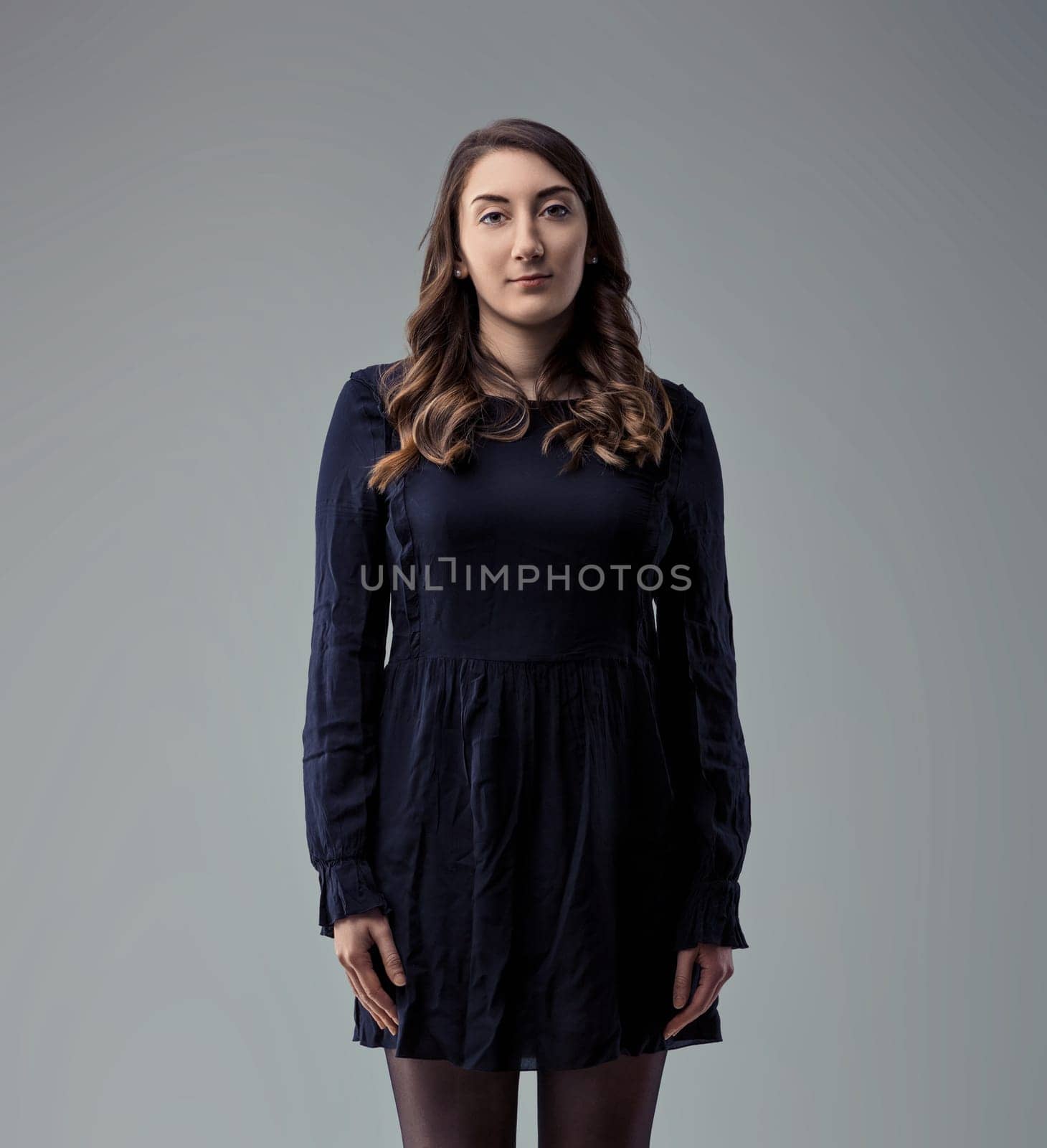 Young woman with long wavy brown hair, wearing a dark blue dress and black tights, standing against a neutral gray background, looking directly at the camera with a serious and confident expression