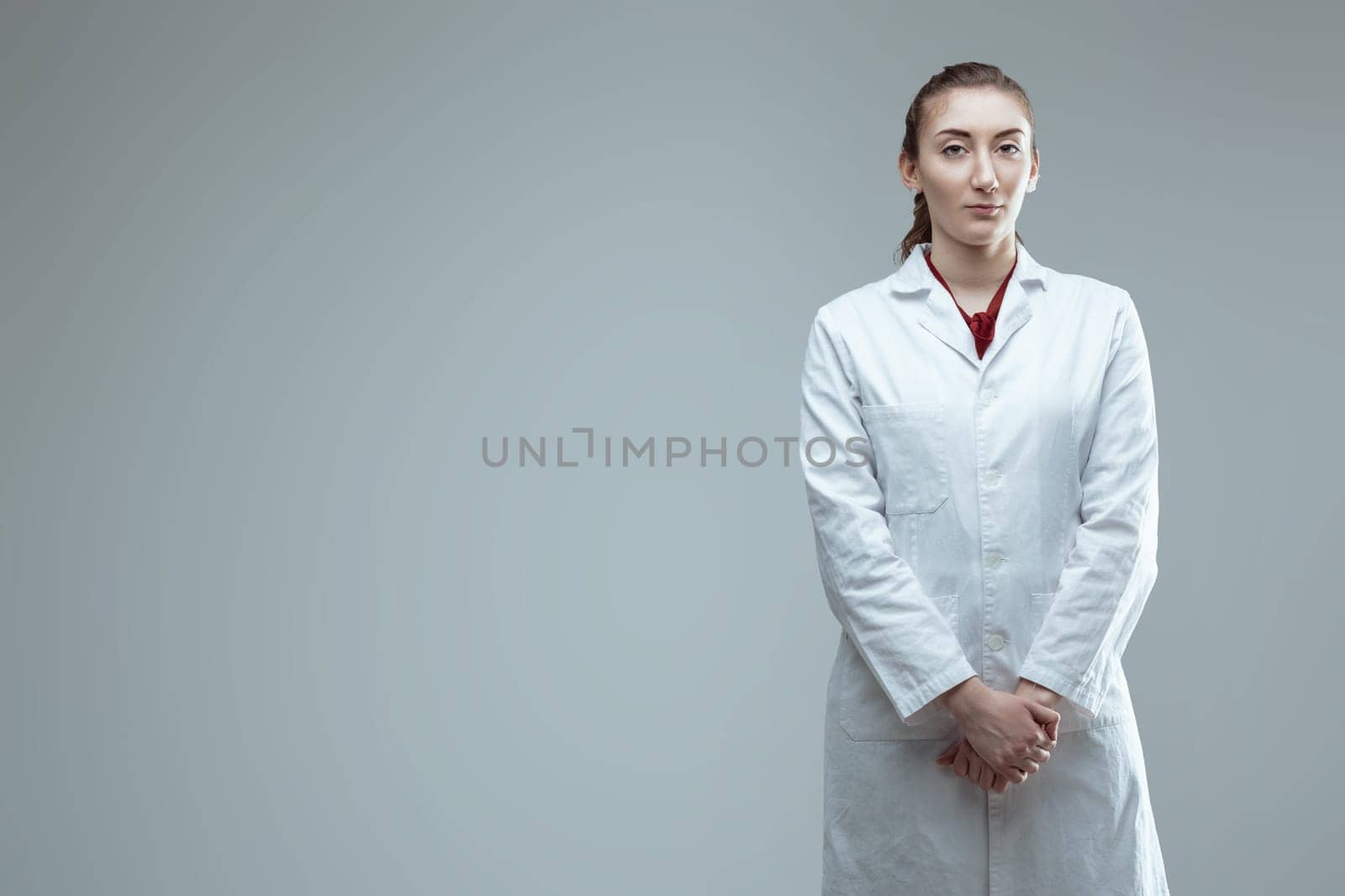 Young confident female technician wearing lab coat is posing in a studio with a serious facial expression, looking directly at the camera with her hands clasped in front of her