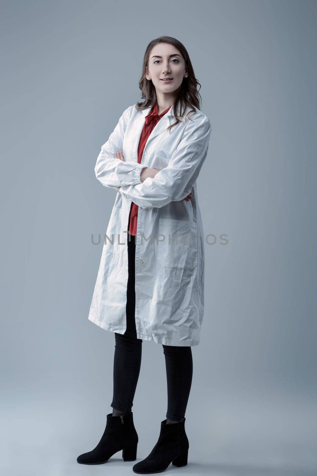 Young confident female technician wearing a lab coat poses with crossed arms in a studio, looking directly at the camera with a serious, determined expression