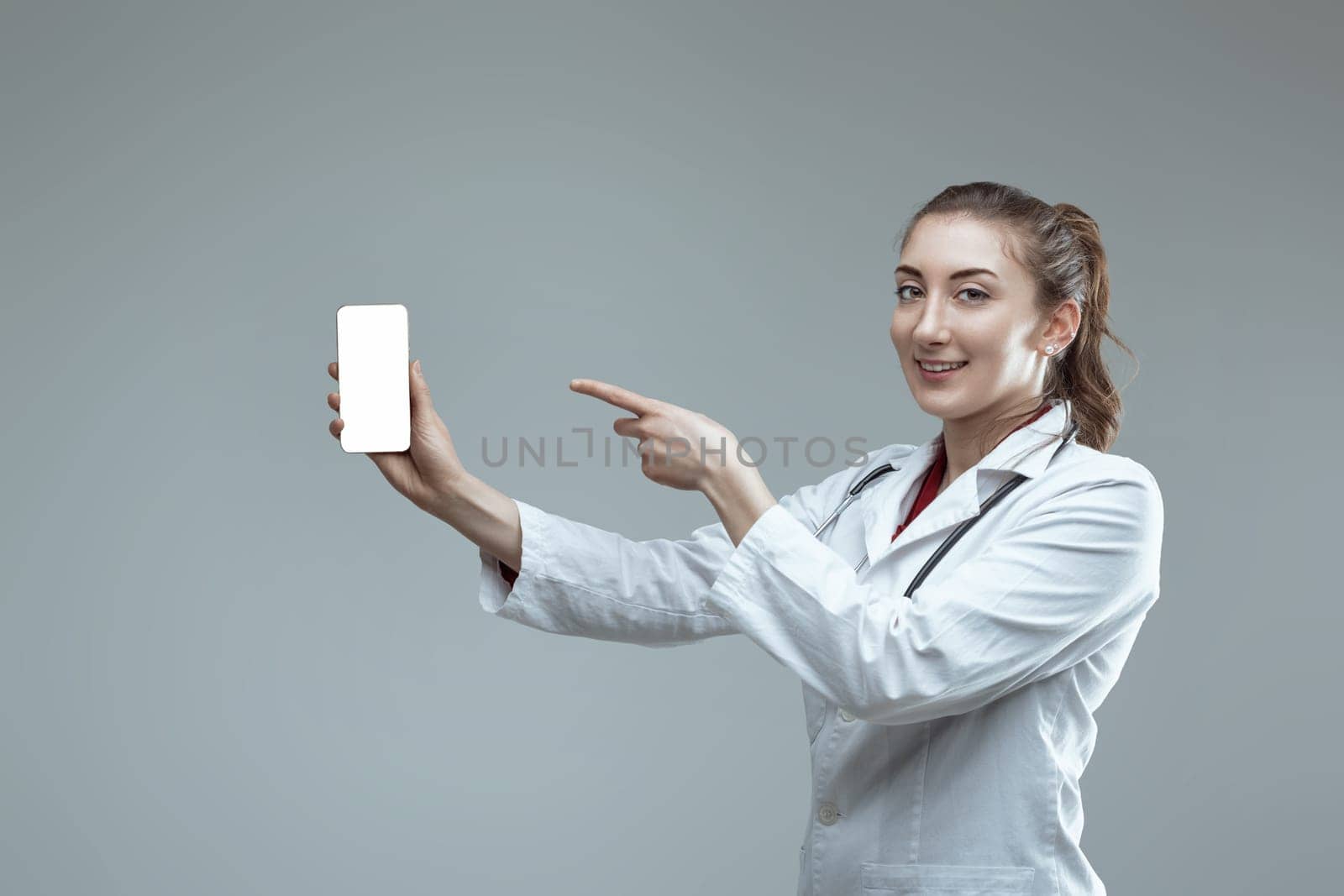 Young female doctor wearing white coat and stethoscope is showing mobile phone with blank screen and pointing at it with index finger, standing against grey background