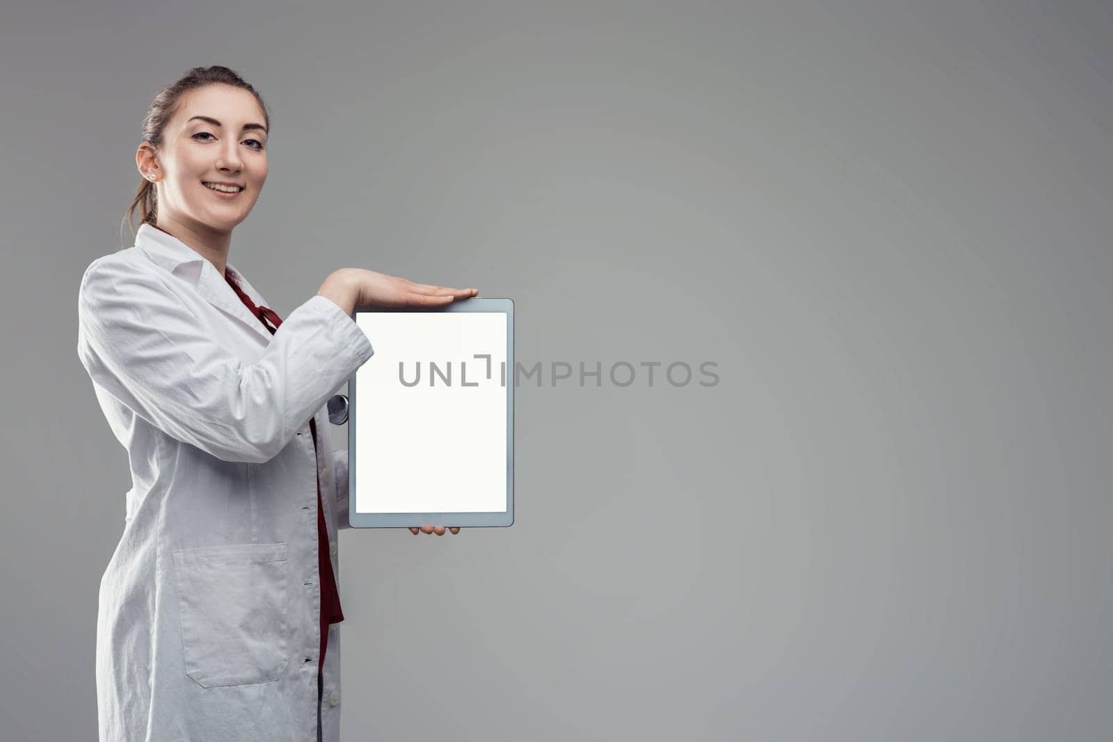 Young female doctor wearing lab coat and stethoscope is showing a digital tablet with blank white screen while standing on a gray background