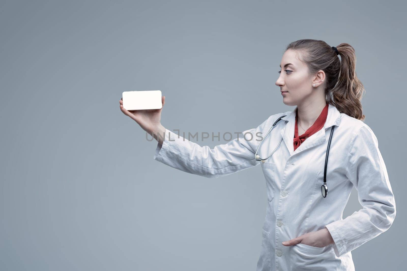 Young female doctor wearing a lab coat and stethoscope, presenting a smartphone with a blank white screen, highlighting a medical app or website for patient engagement and telehealth services