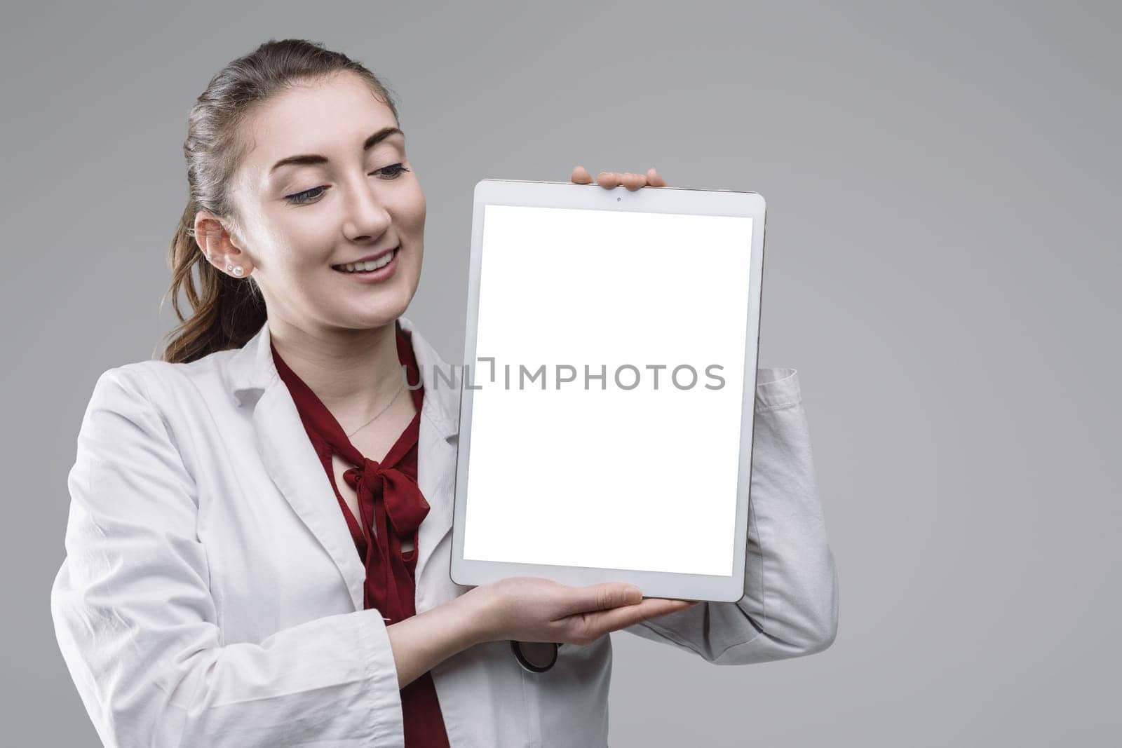 Young female doctor wearing lab coat and stethoscope holding digital tablet with blank screen, looking at it and smiling on plain background