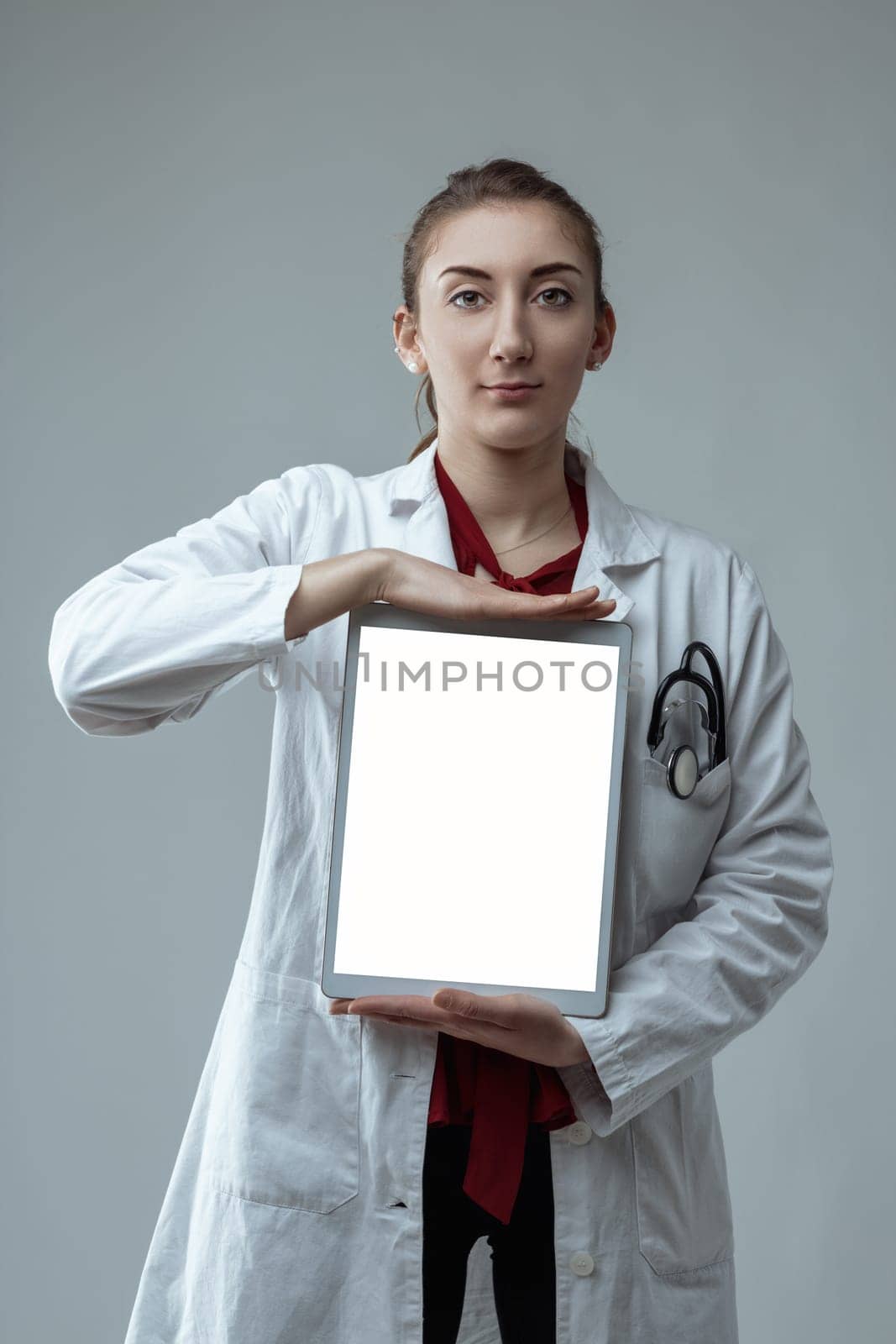 Young female doctor wearing a white coat and stethoscope is holding a digital tablet with a blank screen, providing ample space for custom content related to healthcare and technology