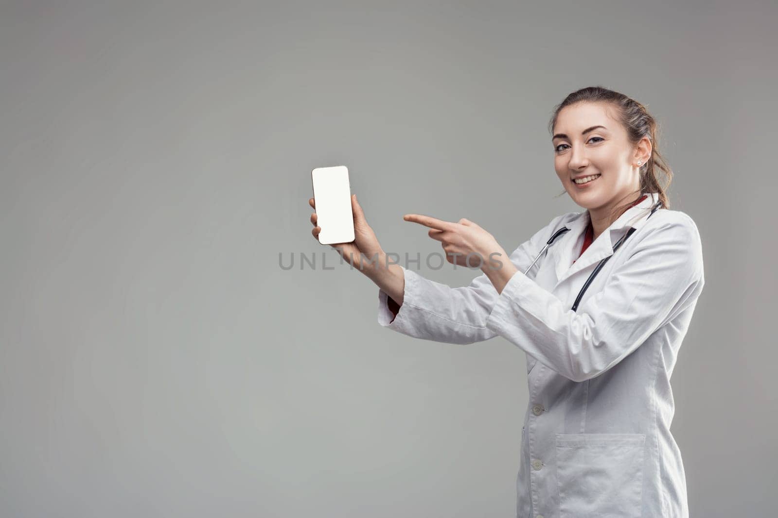 Young female doctor wearing lab coat and stethoscope is showing mobile phone with blank white screen and pointing at it with index finger. Recommending medical app. Standing on plain gray background