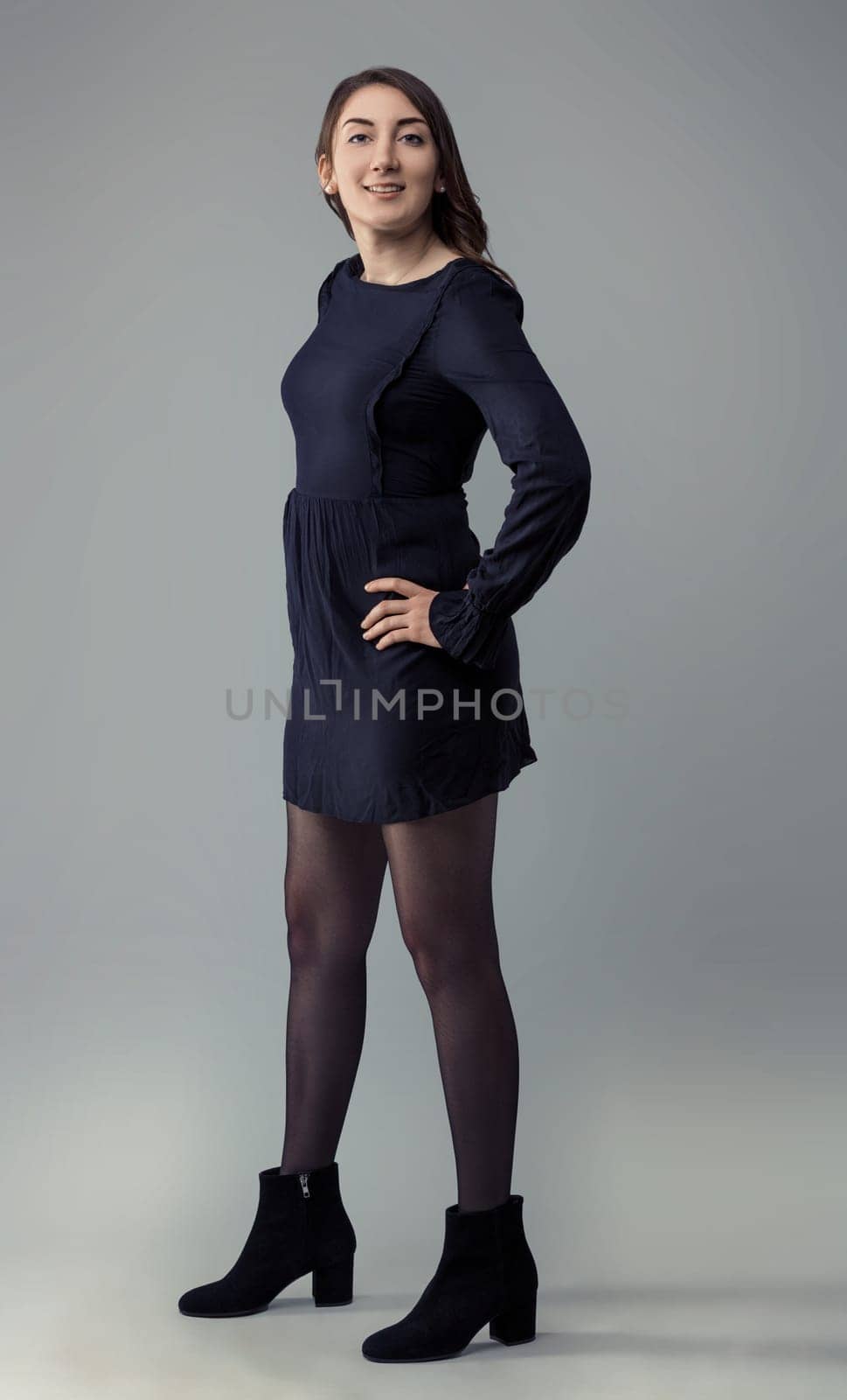 Young woman in a dark dress and ankle boots smiles confidently with her hand on her hip, exuding a sense of self-assurance and empowerment