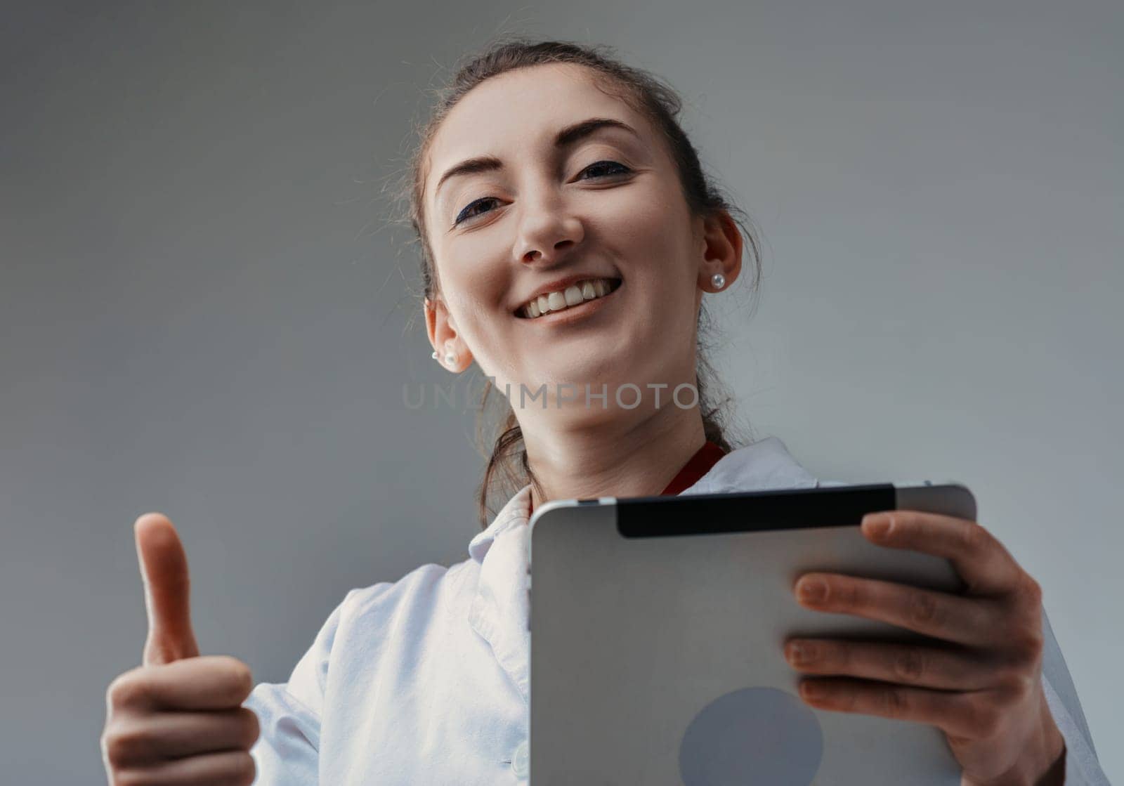 Young female technician or engineer wearing a lab coat smiles and shows a thumbs up gesture while holding a tablet in front of a gray background