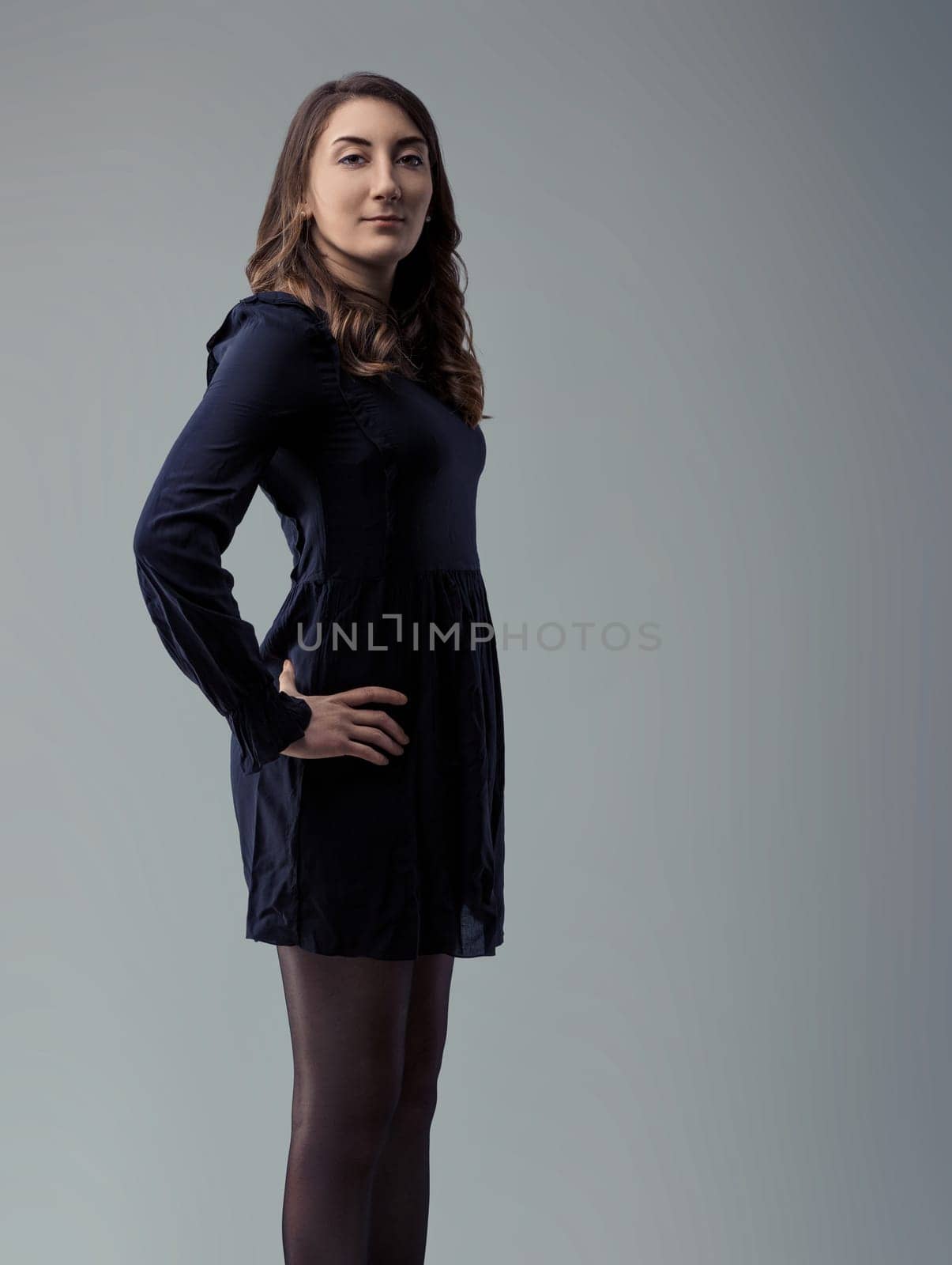 Young woman with long brown hair, wearing a dark dress and tights, stands in profile against a gray backdrop, her hand resting confidently on her hip while looking directly at the camera