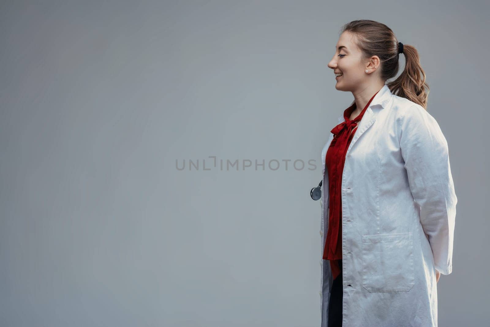 Side view of a young female doctor wearing a white coat and stethoscope, standing with her hands behind her back and smiling warmly, conveying confidence and professionalism in healthcare