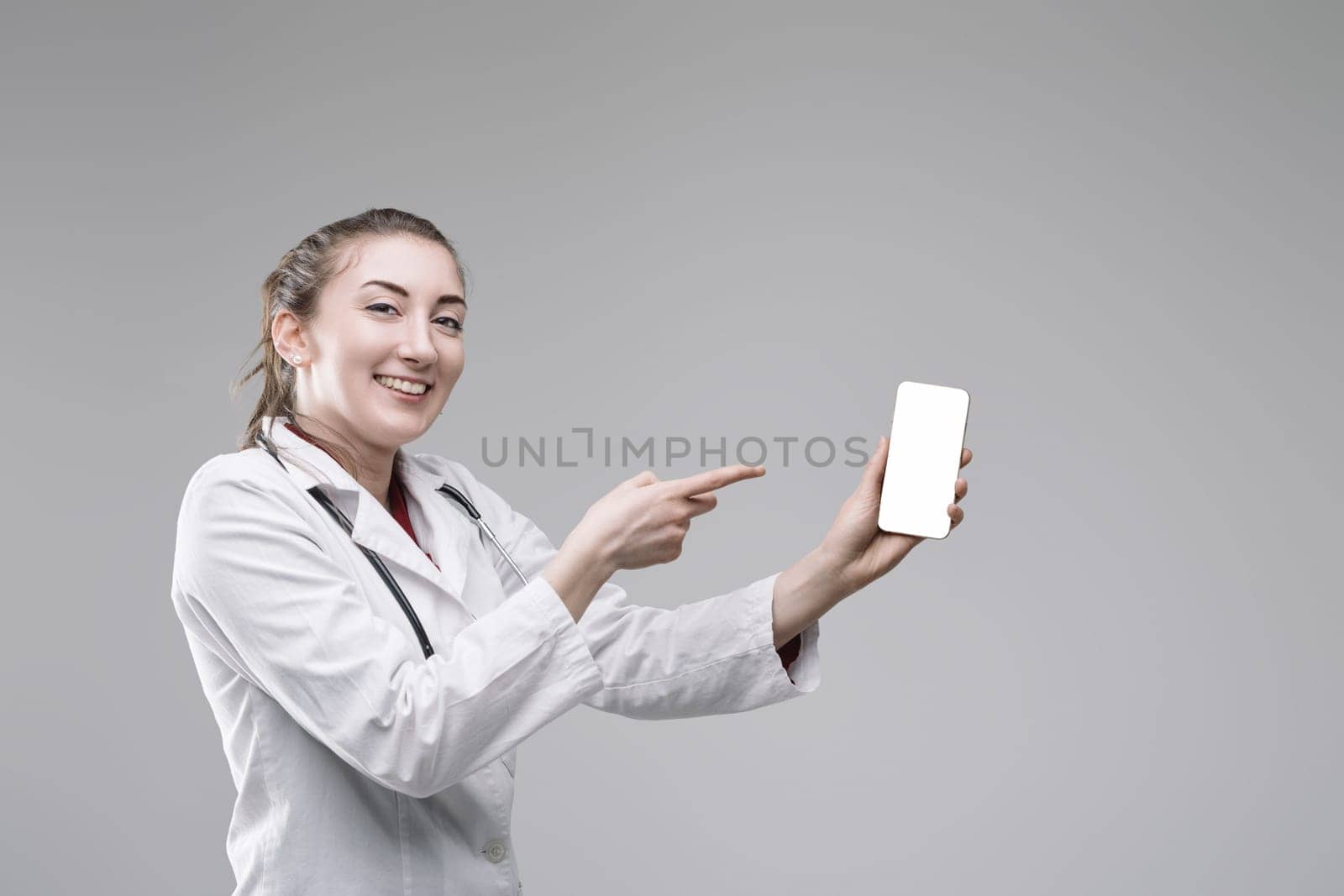 Young female doctor wearing lab coat and stethoscope is holding smartphone with blank screen and pointing at it with index finger, recommending medical app or telemedicine services