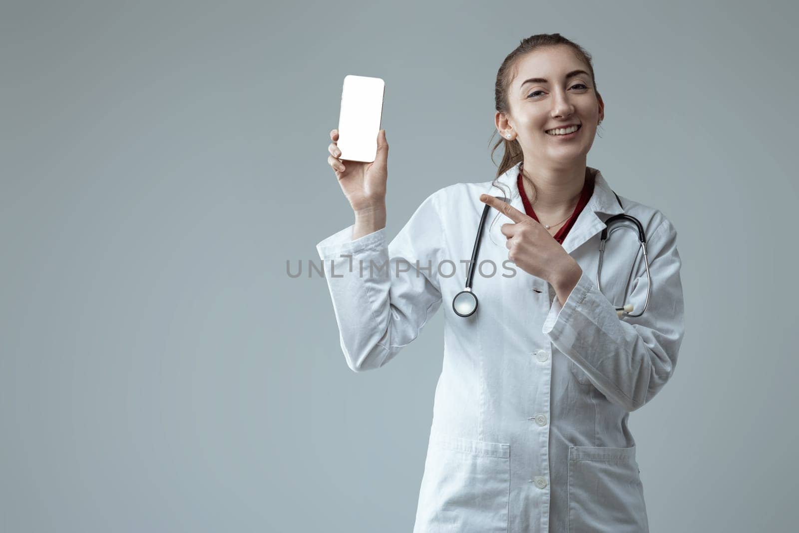 Young female doctor wearing lab coat and stethoscope is holding smartphone with white display and pointing at it with index finger while smiling at camera on gray background