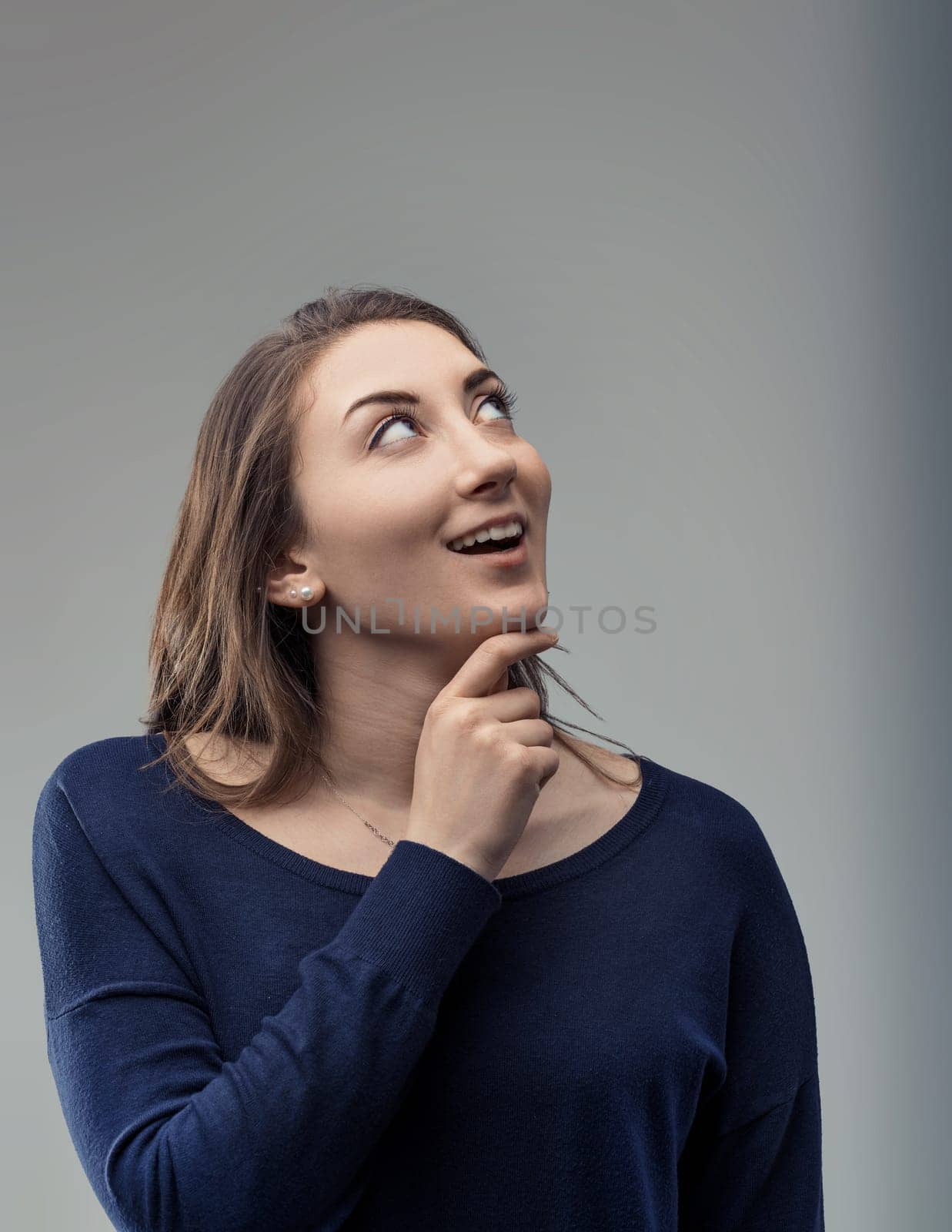 Pensive young woman gazing upwards with a smile, resting her hand on her chin, contemplating future projects and feeling a surge of inspiration, hope, and ambition