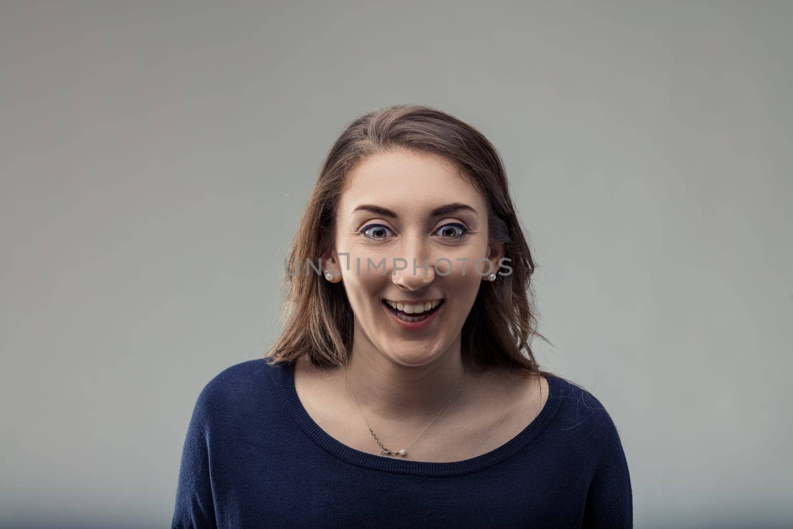 Surprised young woman gasping and smiling with joy, radiating excitement and happiness against a light gray background, showcasing her cheerful expression and vibrant energy