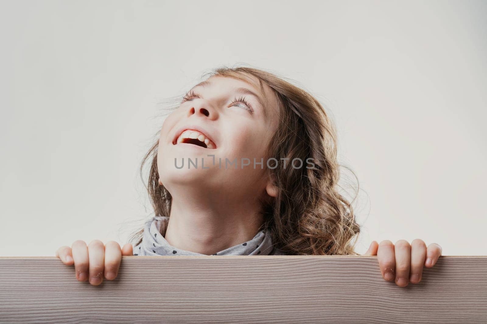 Portrait of a cheerful girl with long brown hair, hiding behind a wooden board and looking up with a happy expression, showing curiosity and joy