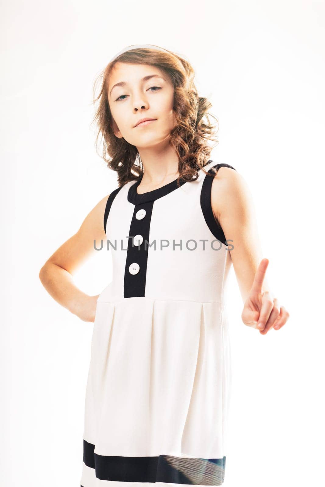 Young girl with curly hair and a stylish dress stands with her hands on her hips. Raising her index finger in a gesture of warning or disapproval. Conveying a sense of seriousness and assertiveness