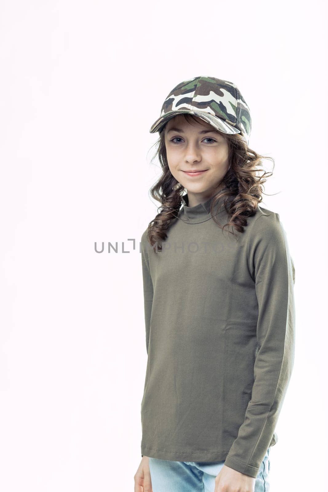 Smiling preteen girl with long brown hair, wearing a camouflage baseball cap, olive green turtleneck, and light blue jeans, posing confidently against a white background