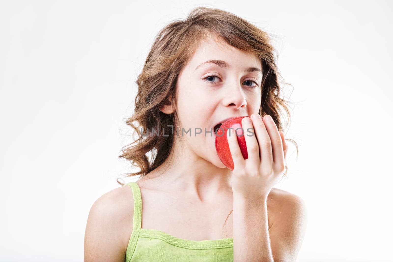 Young girl with wavy brown hair, wearing a light green tank top, takes a large bite out of a crisp, shiny red apple against a clean white backdrop