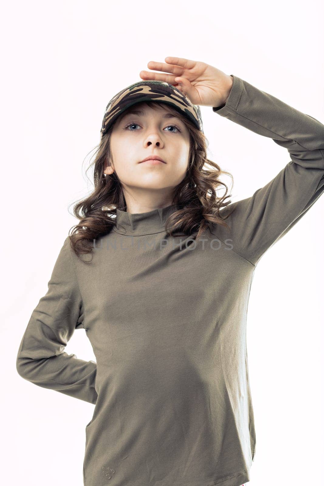 Portrait of a confident young girl standing against a white background, wearing a camouflage cap and saluting with her hand on her forehead, exuding a sense of pride and determination