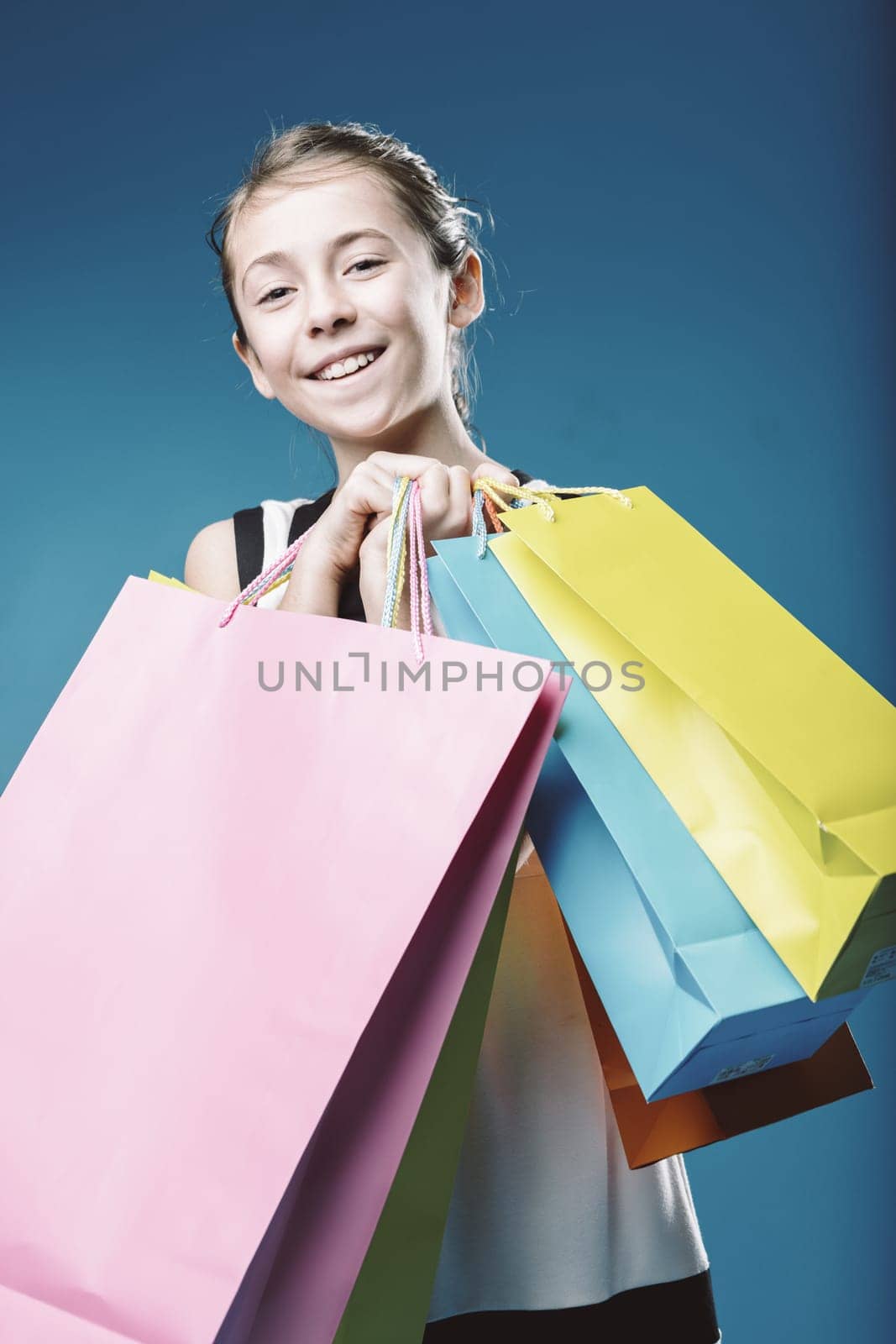 Studio shot of a cheerful young girl holding colorful shopping bags against a vibrant blue background, showcasing the joy and excitement of consumerism and retail therapy