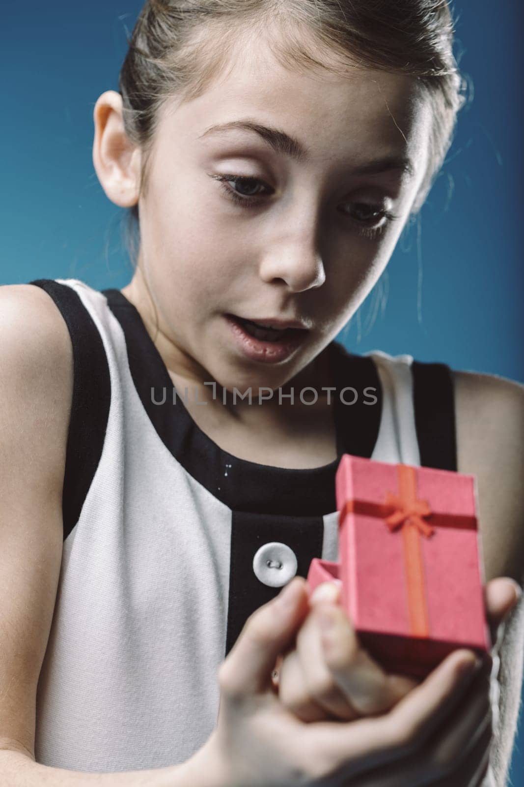 Young girl holds a small, red gift box in her hands, her eyes wide with surprise and curiosity as she opens it against a vibrant blue background