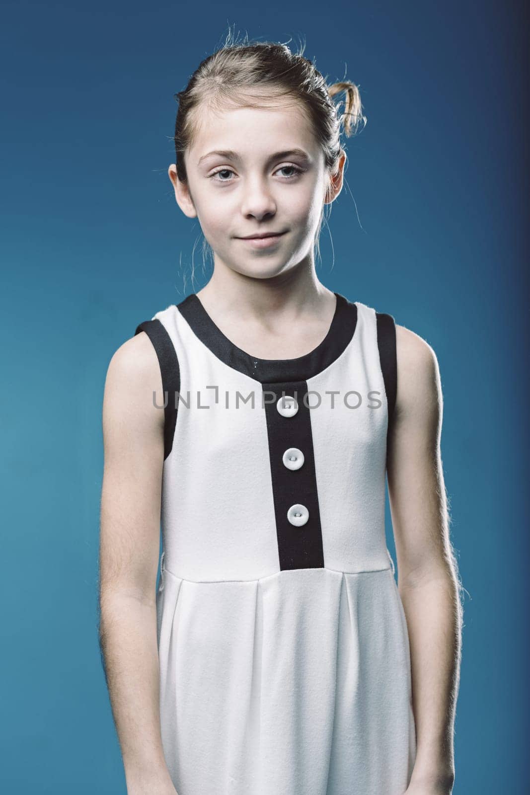 Studio portrait of a smiling young girl wearing a modern white dress with black details, standing against a vibrant blue backdrop, exuding confidence and style