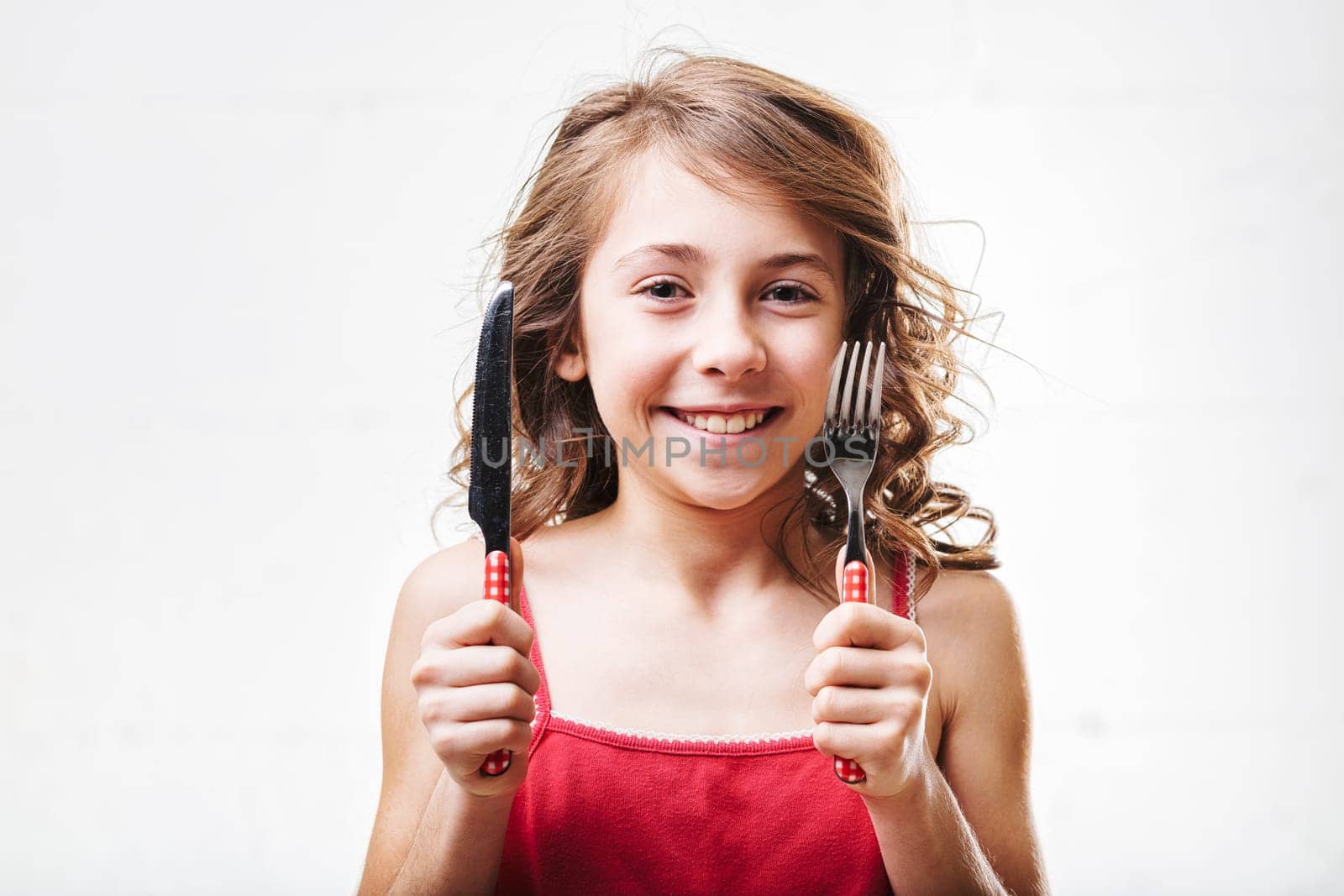 Smiling young girl with wavy hair holds a fork and knife, radiating excitement and joy while eagerly anticipating a delicious meal