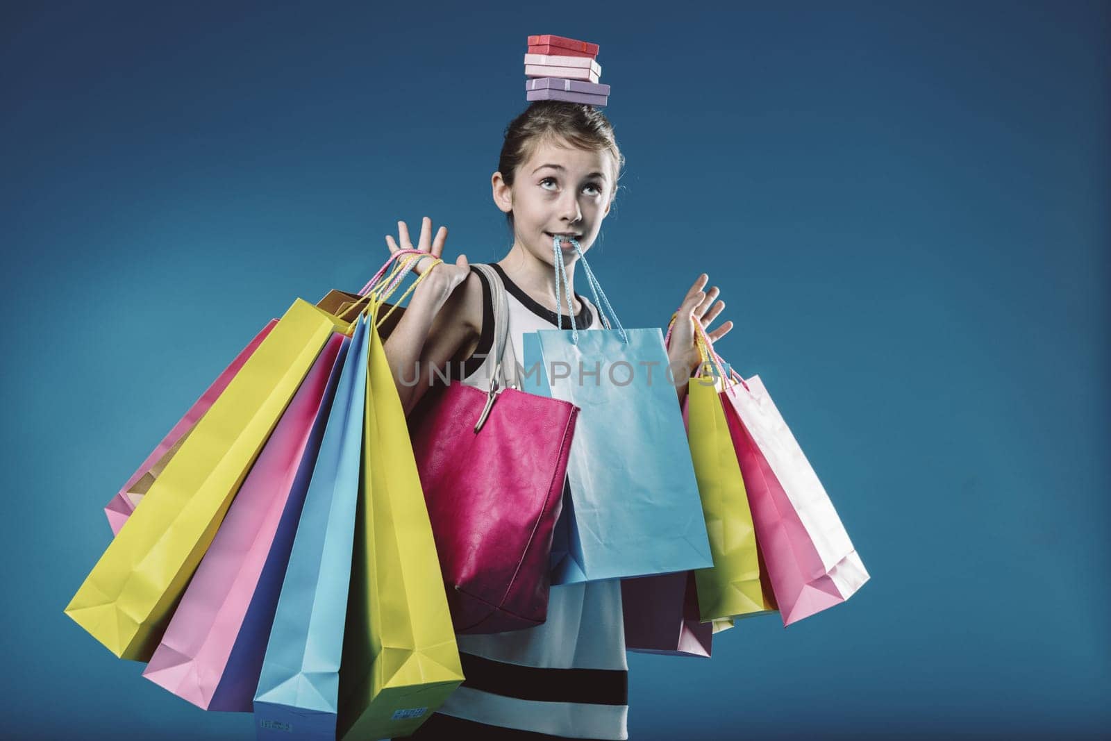 Young girl skillfully balances a stack of colorful gift boxes on her head while juggling numerous shopping bags, radiating happiness and excitement after a successful shopping spree