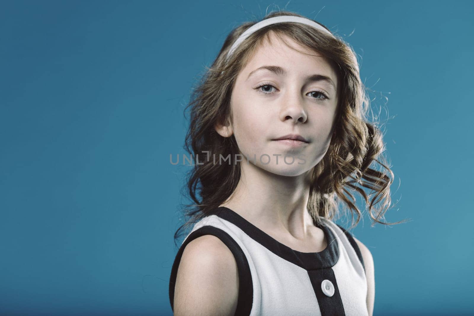 Studio portrait of a smiling young girl with brown curly hair and a white headband, wearing a black and white dress, posing against a vibrant blue backdrop